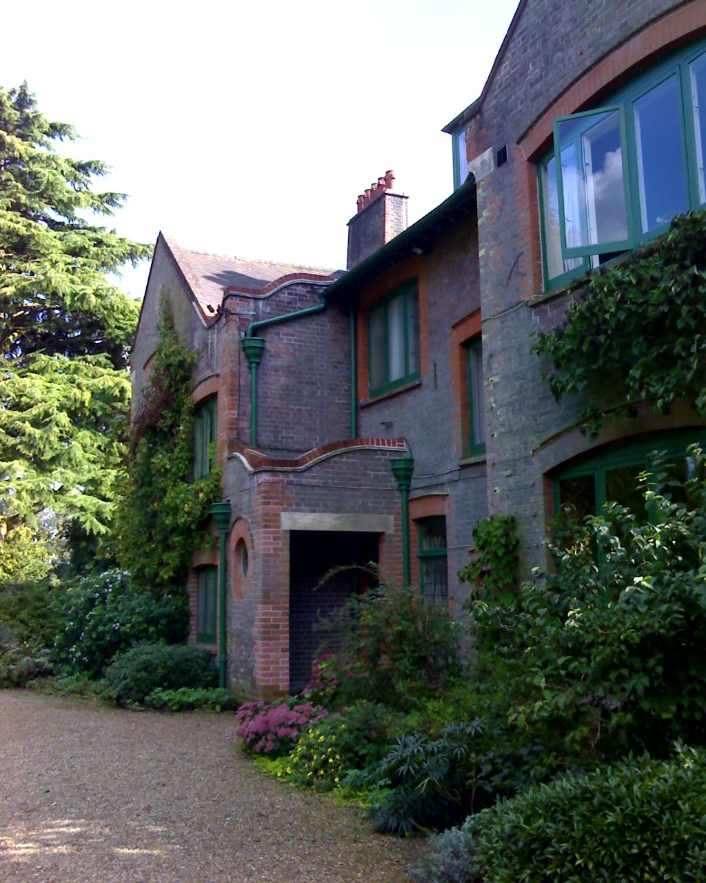 Side of large red brick house surrounded by trees and plants.