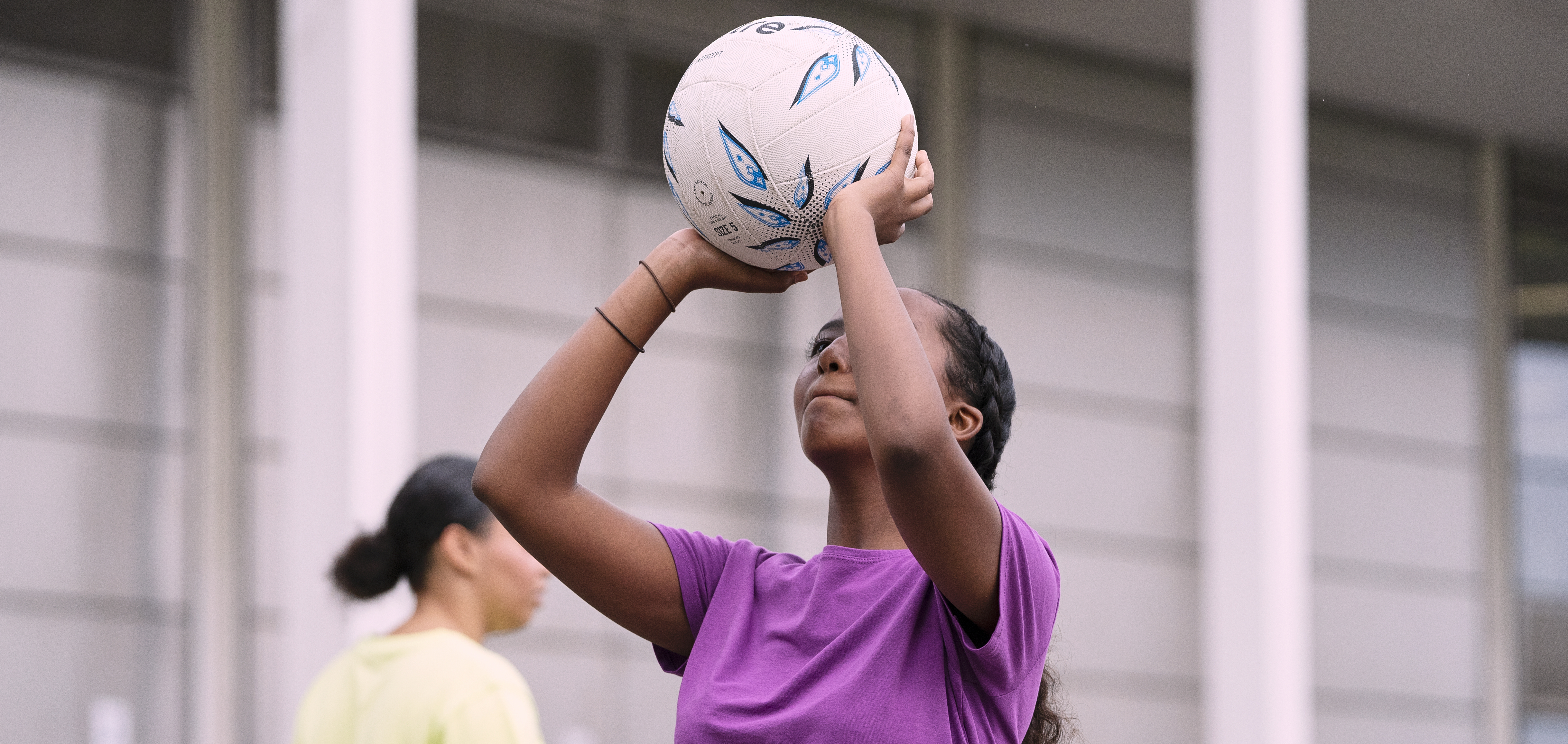 girl playing netball holding the ball above her head wearing a purple t shirt